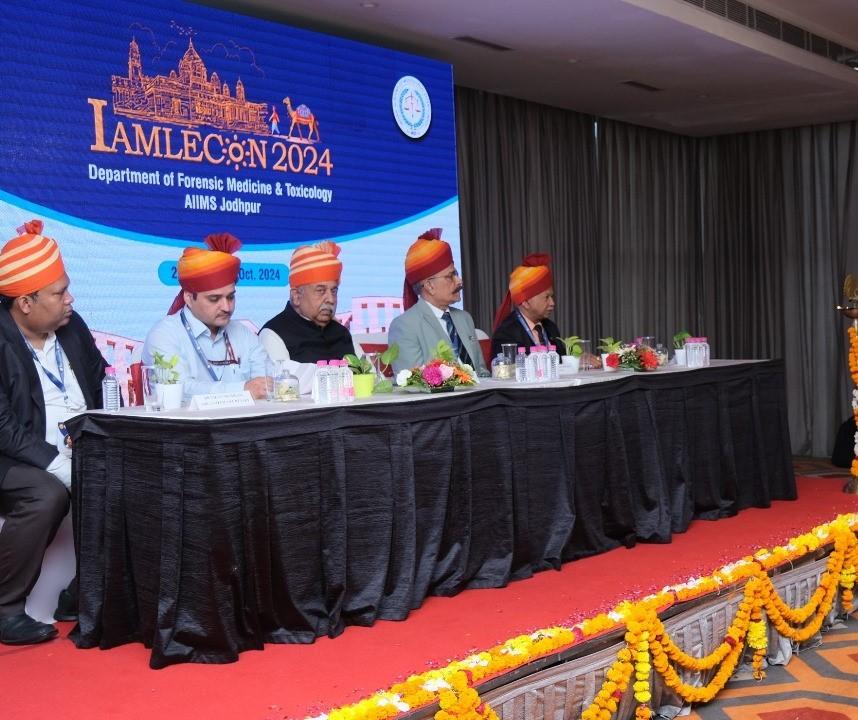  Panelists at a conference wearing traditional turbans behind a table decorated with flowers.